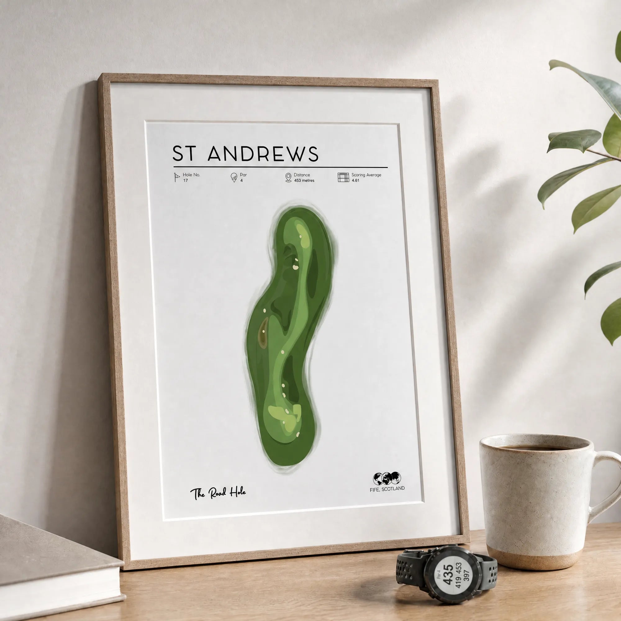 Golf wall art leaning on light timber desk, framed St Andrews golf course print, ceramic coffee cup, soft morning light